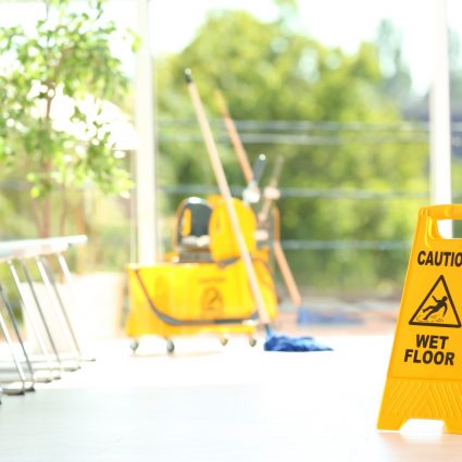 Safety sign with phrase Caution wet floor and blurred mop bucket on background. Cleaning service